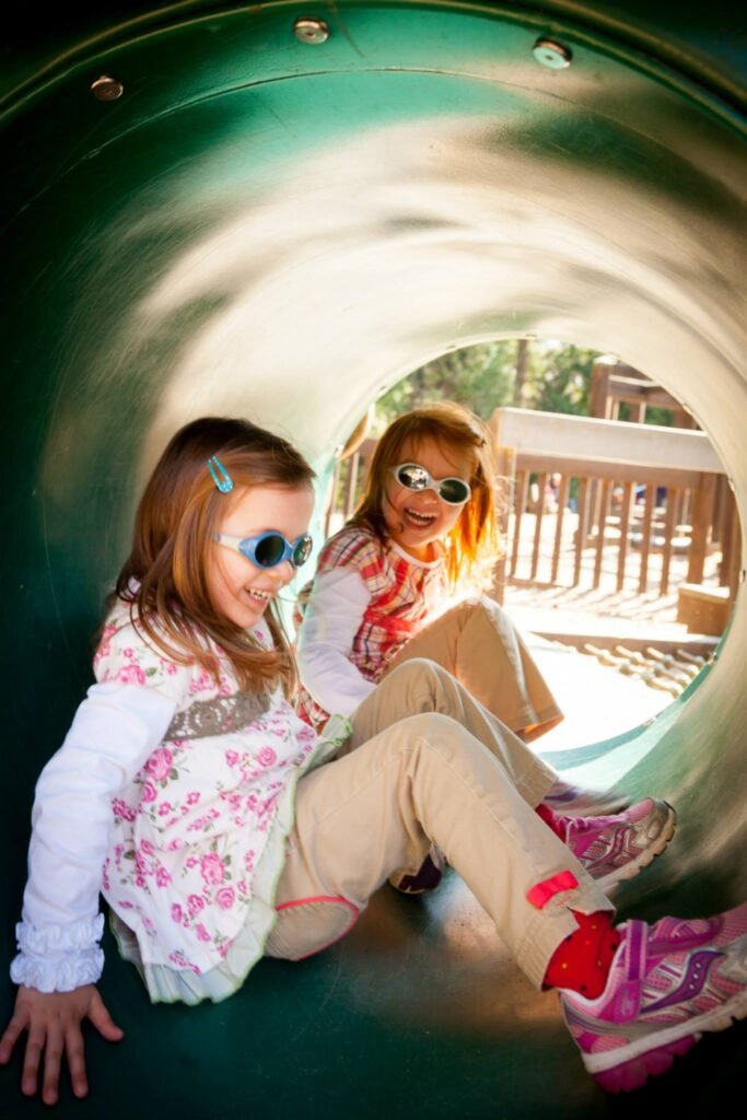 Two children wearing sunglasses sitting inside a green playground tunnel. They appear to be enjoying their time at a playground.