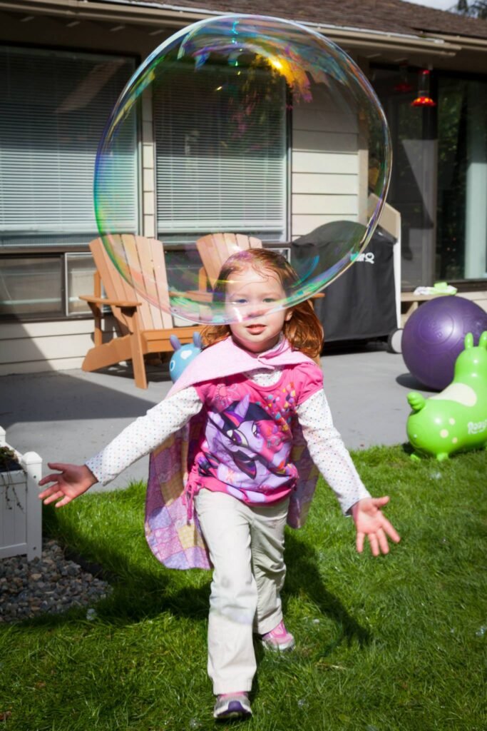 A child runs on a backyard lawn while a giant iridescent bubble floats overhead. The scene shows a sunny day with toys nearby.