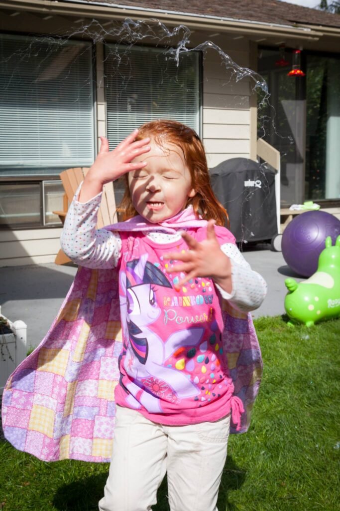 A young child in a pink Minnie Mouse shirt and checkered cape plays outdoors while water sprays behind them.