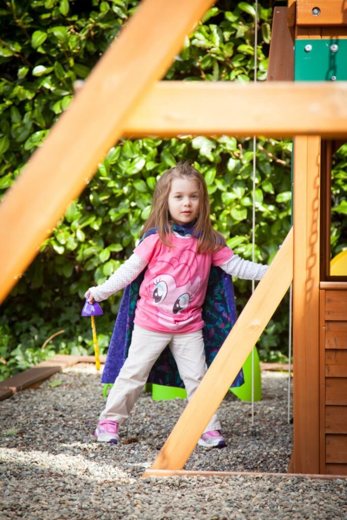 A young child wearing a pink shirt stands on a wooden playset holding a purple toy. Green foliage serves as the background in this backyard scene.