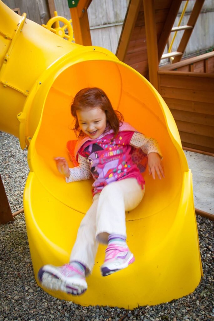 A child in a pink jacket enthusiastically sliding down a large yellow playground tube. The scene captures a moment of pure joy and childhood energy.