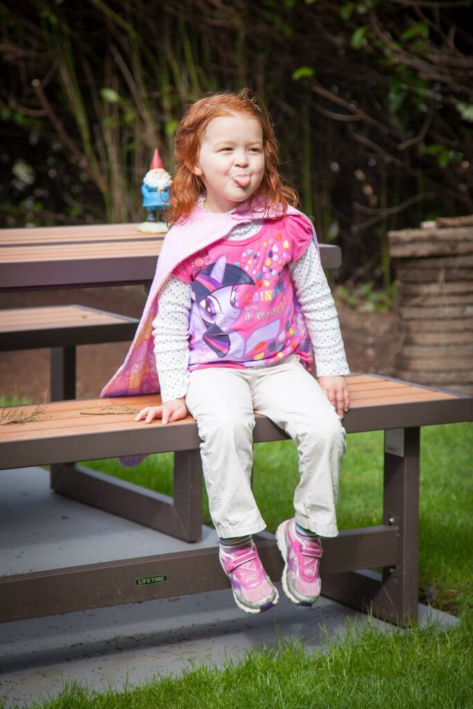 A child in a pink coat sits on a backyard picnic table with a tiny gnome in the background. The photo is taken in a backyard setting.