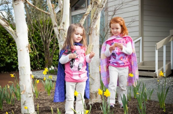 Two children in colorful costumes standing among blooming daffodils and young trees in a Pacific Northwest garden.