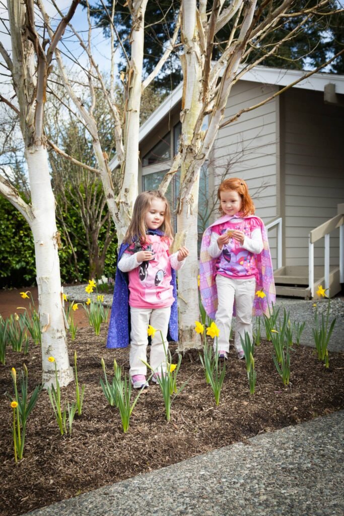 Two children in colorful costumes standing among blooming daffodils and young trees in a Pacific Northwest garden.