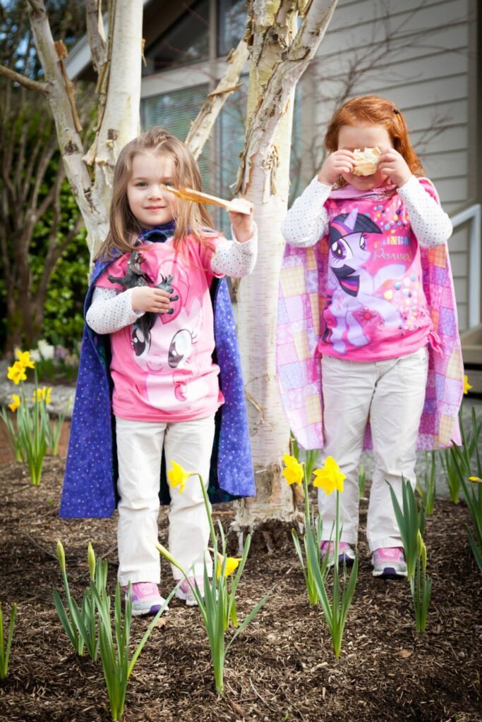 Two children in pink dresses stand among blooming yellow daffodils, playfully eating sweets on sticks in a garden setting.