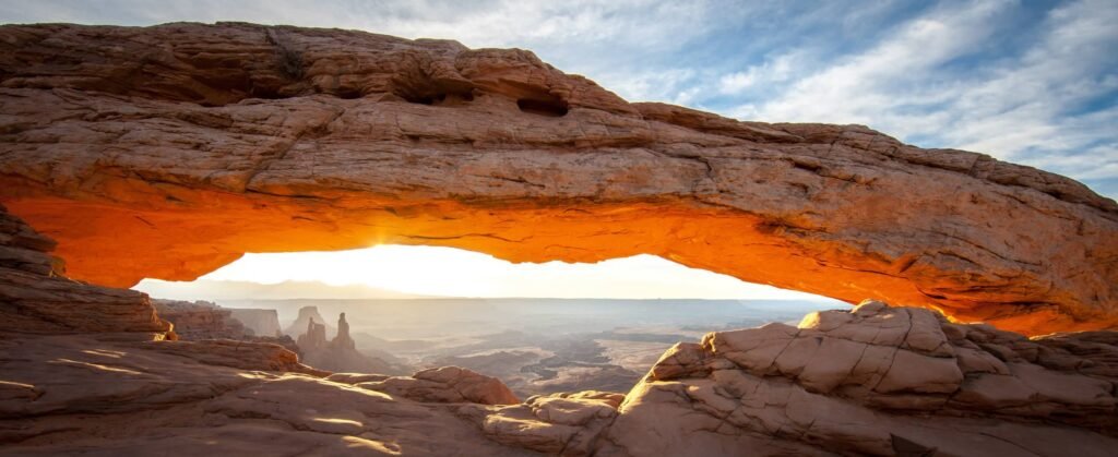 A large rock arch with orange light shining through at sunset. The landscape in the distance is visible under the arch.