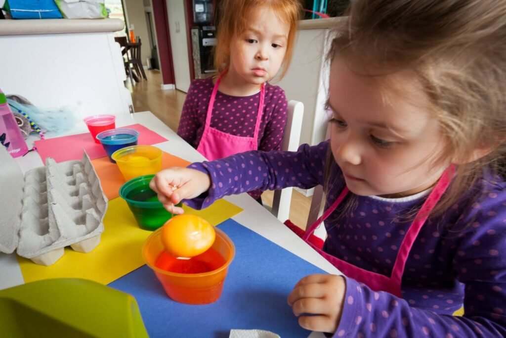Children wearing aprons dyeing an egg in a bowl of red liquid on a colorful table.