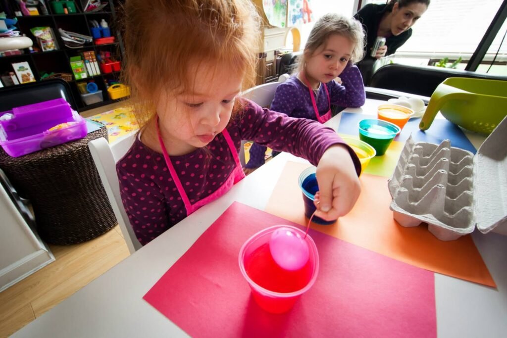 Young children wearing aprons participate in a hands-on activity, carefully pouring liquid into a cup with an egg carton nearby.