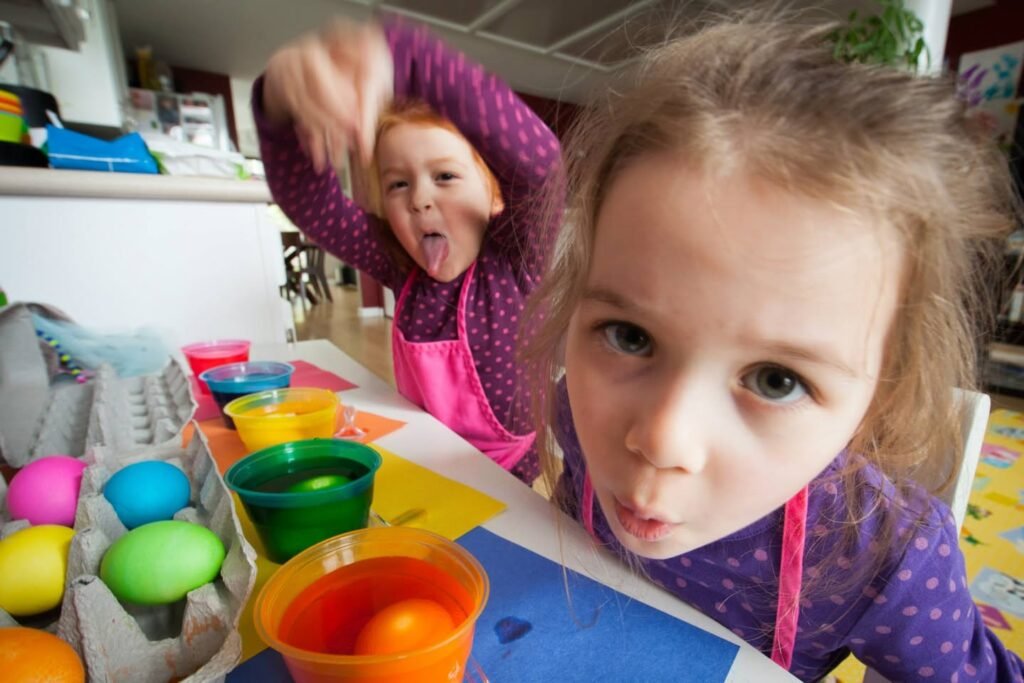 Two children in purple shirts enjoy a creative egg-painting activity with colorful bowls and egg cartons on a table.