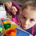 Two children in purple shirts enjoy a creative egg-painting activity with colorful bowls and egg cartons on a table.
