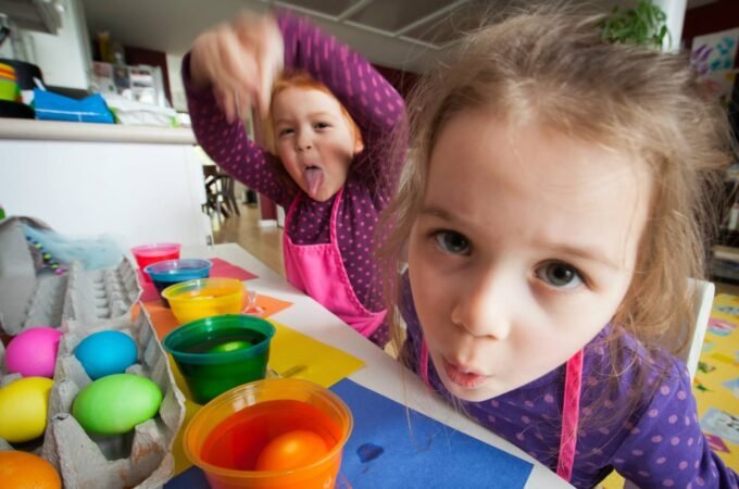 Two children in purple shirts enjoy a creative egg-painting activity with colorful bowls and egg cartons on a table.