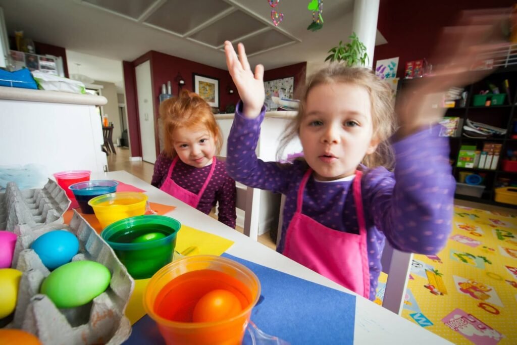 Two children dyeing brightly colored Easter eggs at a kitchen table with paint cups and egg cartons.