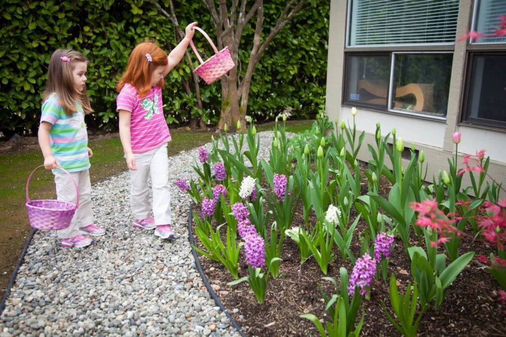 Two children with pink baskets enjoying a garden walk among purple hyacinths and green foliage.