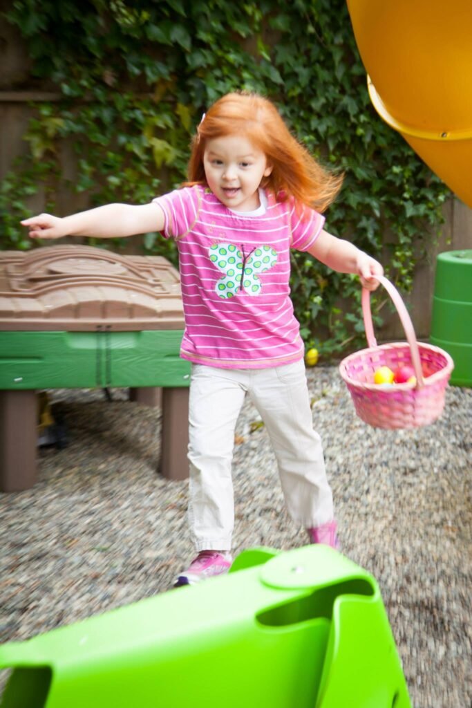 A child in a pink striped shirt runs through a outdoor play area carrying a pink basket of plastic Easter eggs.