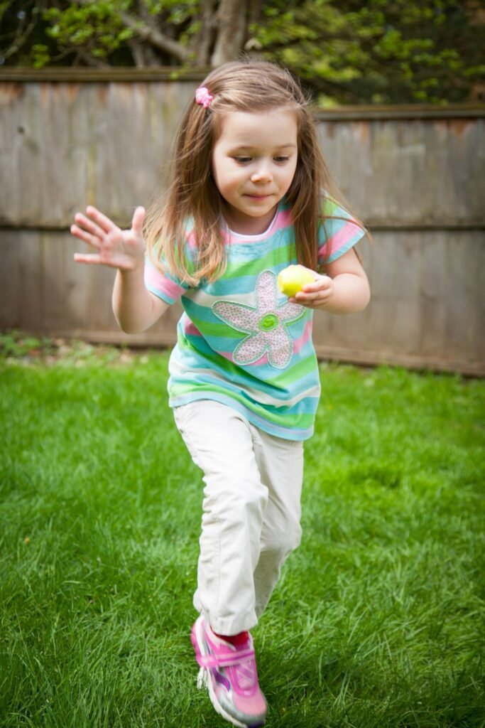 A child runs across a grassy lawn holding a yellow ball, wearing a striped shirt and pink shoes near a wooden fence.