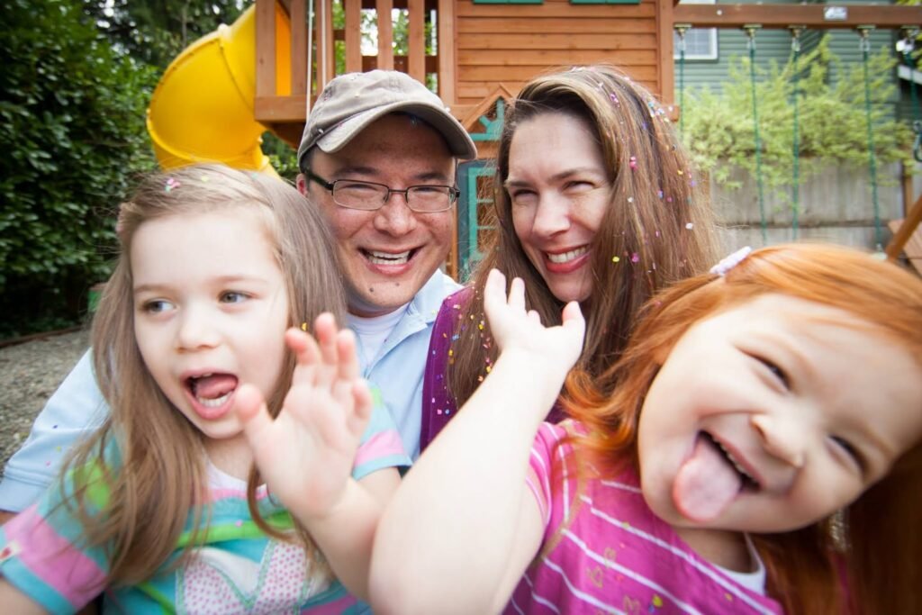 A joyful family of four poses playfully outdoors with a child making a silly face on a yellow playground slide.