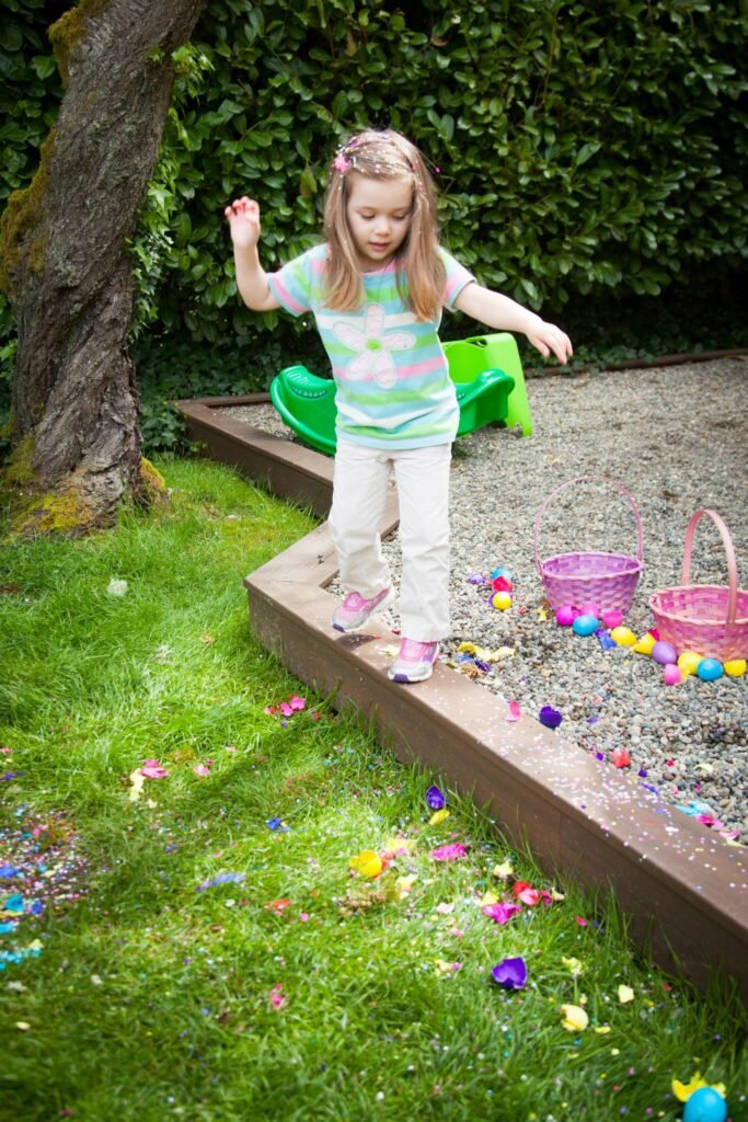 A child stands on a garden ledge near a tree with colorful Easter eggs scattered around.