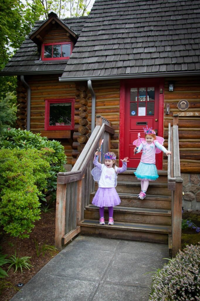 Children in fairy costumes stand on the wooden steps of a rustic log cabin with a bright red front door and cedar shingle roof.