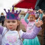 Children wearing purple and pink princess crowns pose joyfully at a theme park event. One child strikes a playful pose while another smiles brightly in the background.