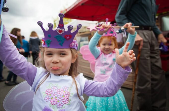 Children wearing purple and pink princess crowns pose joyfully at a theme park event. One child strikes a playful pose while another smiles brightly in the background.