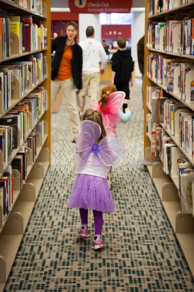 Two children dressed as fairies walking through a bookstore aisle. Tall bookshelves line the patterned tile floor.