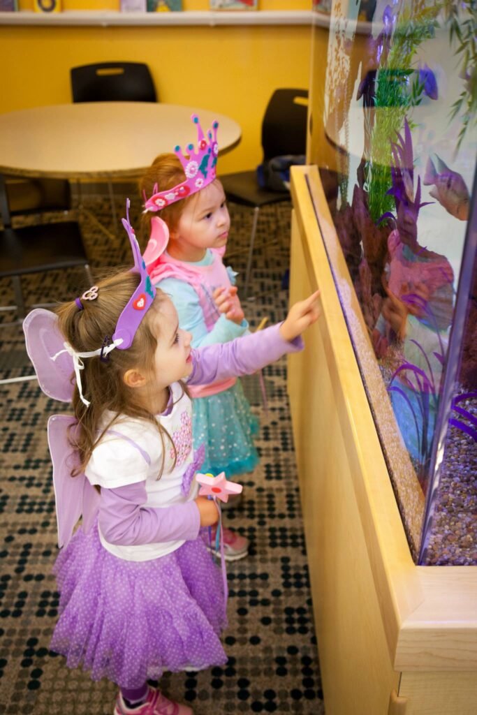 Two children in princess costumes look closely at a large aquarium in a school setting. The colorful fish tank captures their attention as they observe the underwater life.