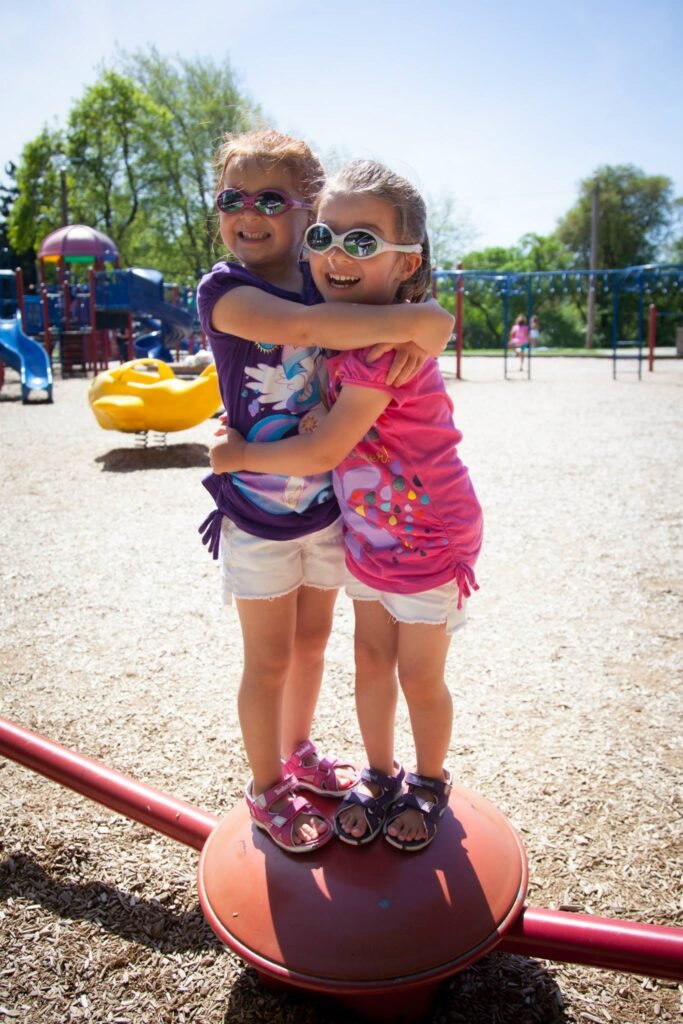 Two children wearing sunglasses hug on a red playground seesaw under a clear blue sky.
