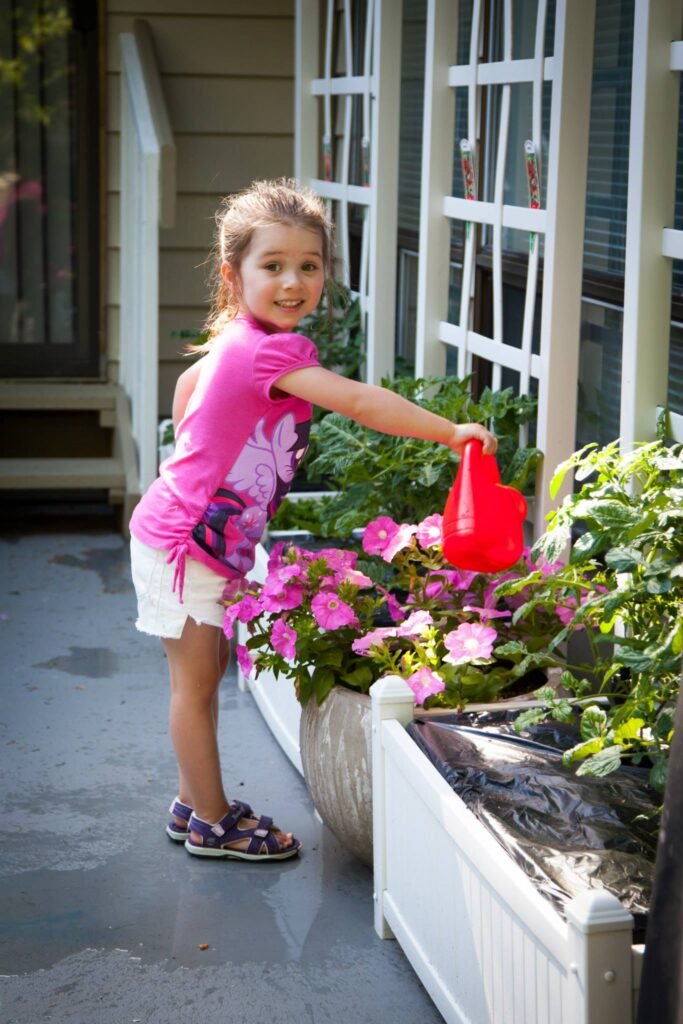 A child waters flowers in a container outside a house. Pink flowers bloom in white planters.