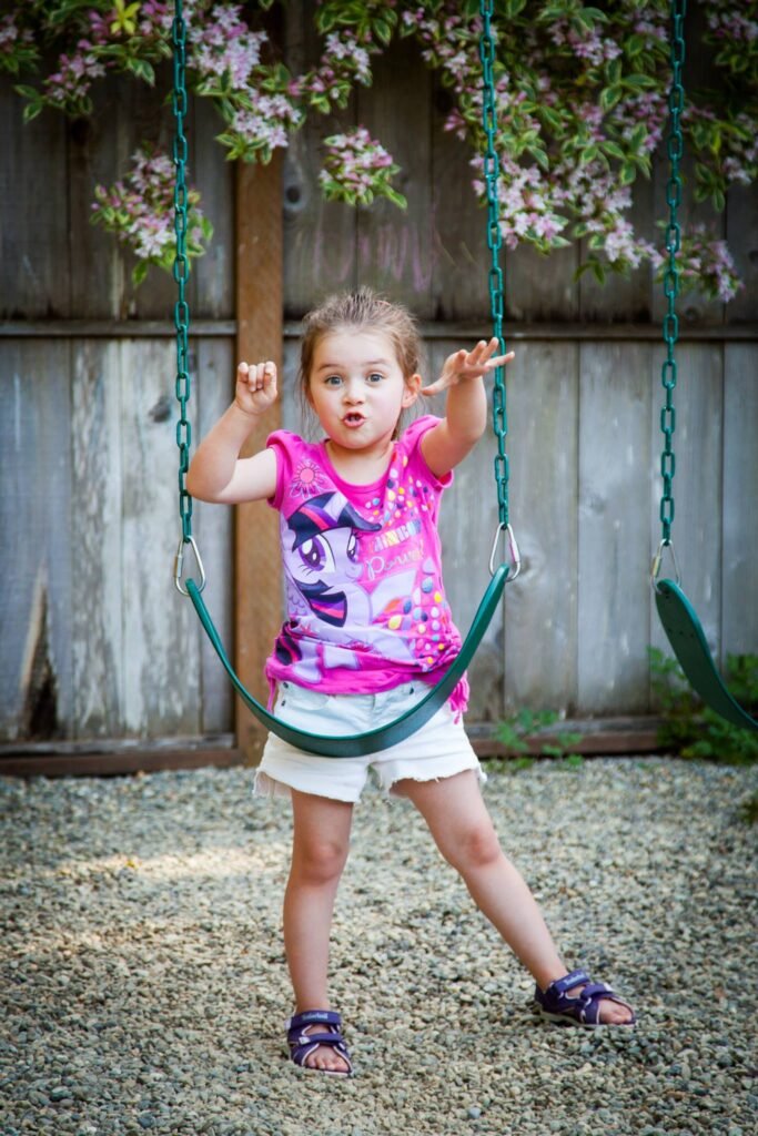 A child stands on gravel beside a green swing, holding the chains in a backyard.