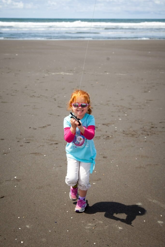A red-haired child wearing sunglasses and a bright blue shirt walks across the wet sand of a beach. The Pacific Ocean waves can be seen in the background.