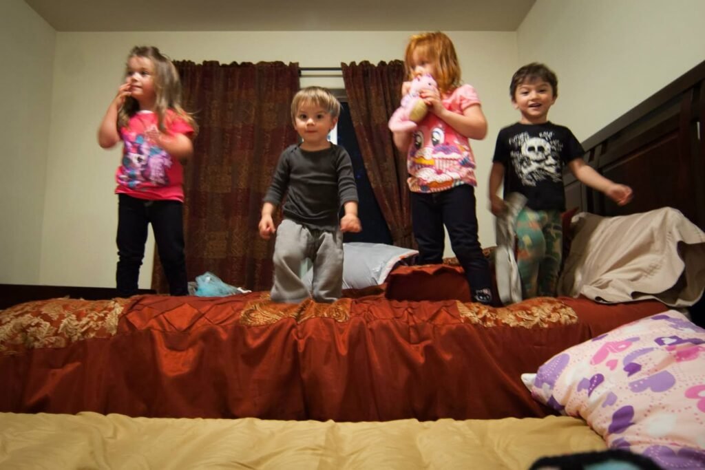 Four children stand on a bed with reddish bedding in a home bedroom.