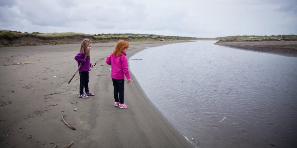 Two children in colorful jackets stand by a calm river mouth on a vast sandy shore, one holding a stick.