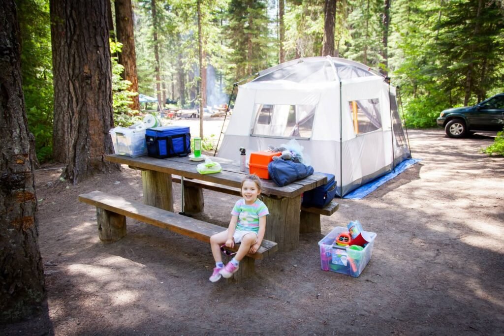A family tent is set up in a sun-dappled forest clearing, next to a wooden picnic table where a child sits.