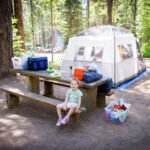 A family tent is set up in a sun-dappled forest clearing, next to a wooden picnic table where a child sits.