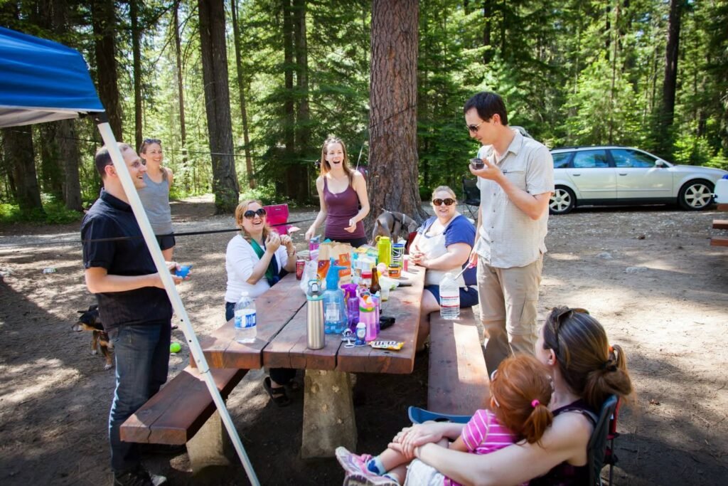A group of people enjoying a picnic at a wooden table in a sun-dappled forest campsite with a blue tarp and car in the background.