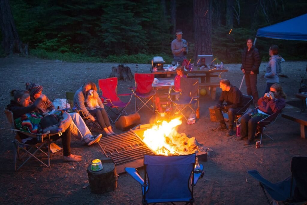 A group of people is gathered around a bright campfire at a nighttime campsite with a blue tent and forest nearby.