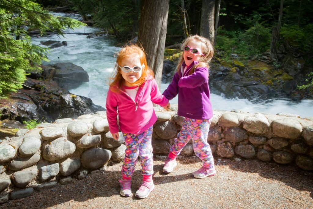Two young children wearing sunglasses and bright jackets play by a rocky stream wall. The rushing water and surrounding greenery create a natural, outdoor picnic spot.