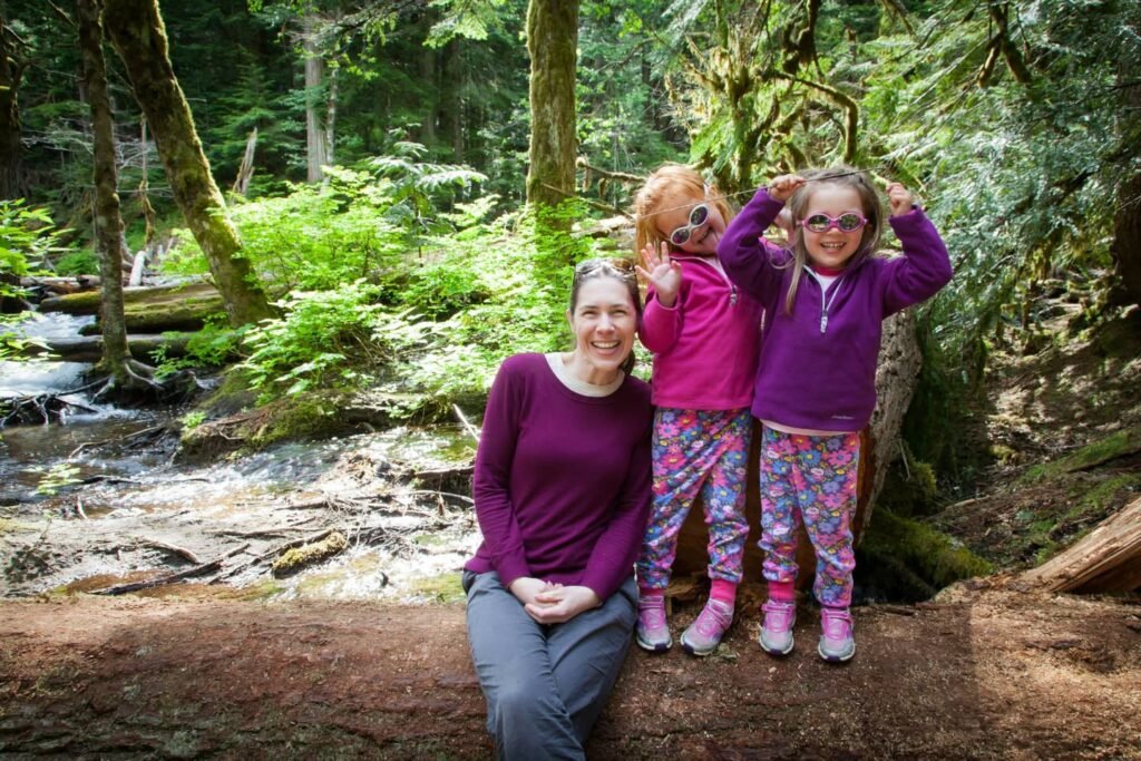A woman and two children sit and stand on a large fallen tree in a lush Pacific Northwest forest with a creek nearby.