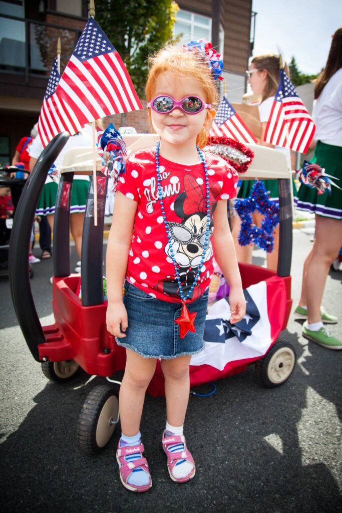 A person in red, white, and blue parade attire stands beside a decorated wagon with American flags.