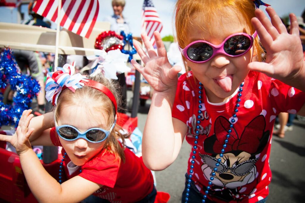 Children waving at the camera during a Fourth of July parade in red, white, and blue outfits.