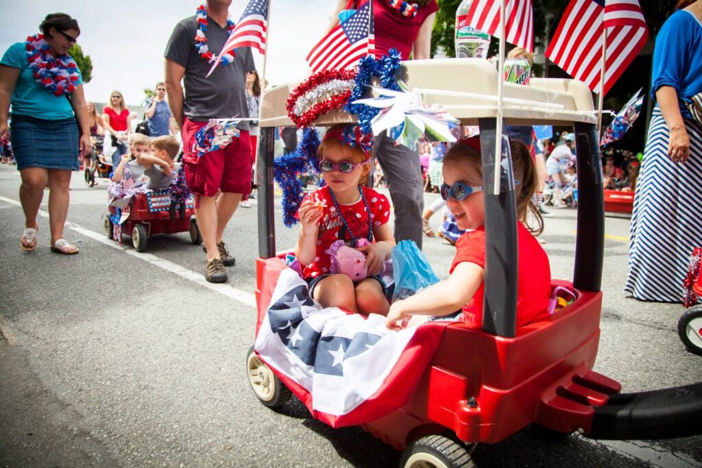 Kids in a red ride-on toy car decorated with an American flag during a parade, with adults walking nearby wearing patriotic leis.