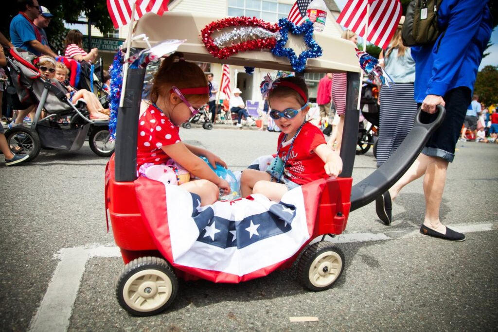 Children sit in a red wagon decorated with American flags and stars during a lively street parade.