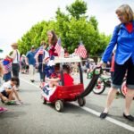 Spectators watch a red flag-decorated wagon in a community parade on a sunny street.