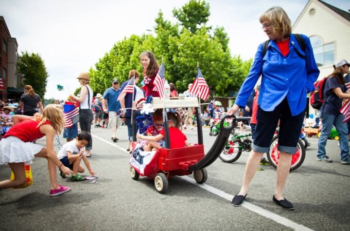 Spectators watch a red flag-decorated wagon in a community parade on a sunny street.