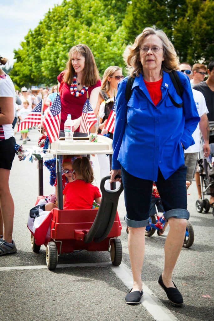 A woman in a blue jacket pulling a red wagon during an outdoor community parade. Spectators line the street holding American flags under sunny skies.