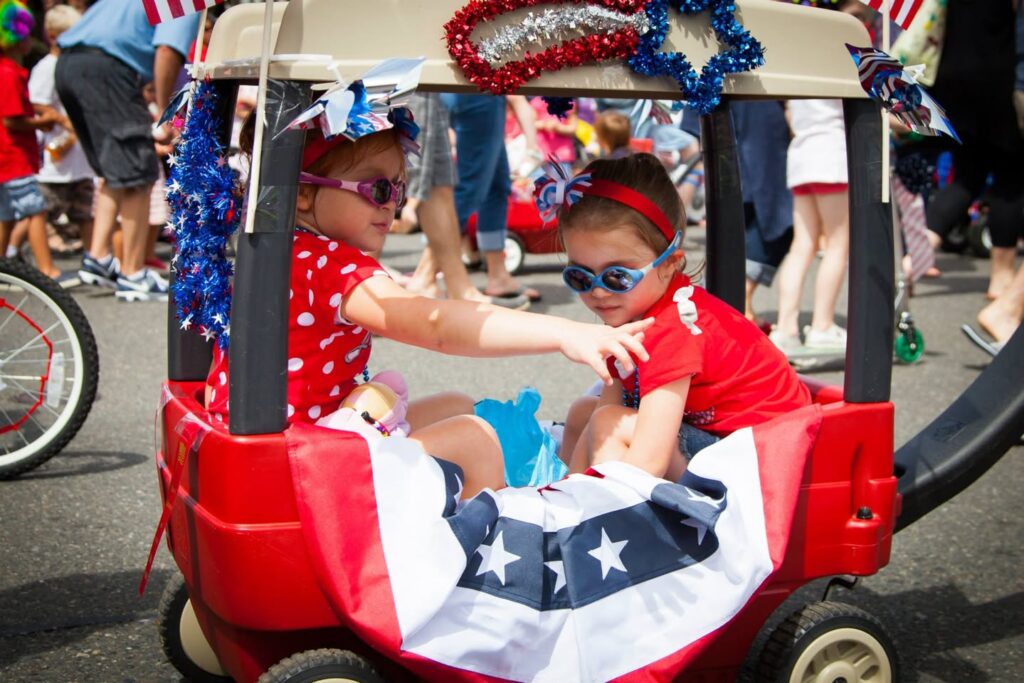 Two children dressed in patriotic red, white, and blue outfits sit in a parade vehicle decorated with American flags and bunting.