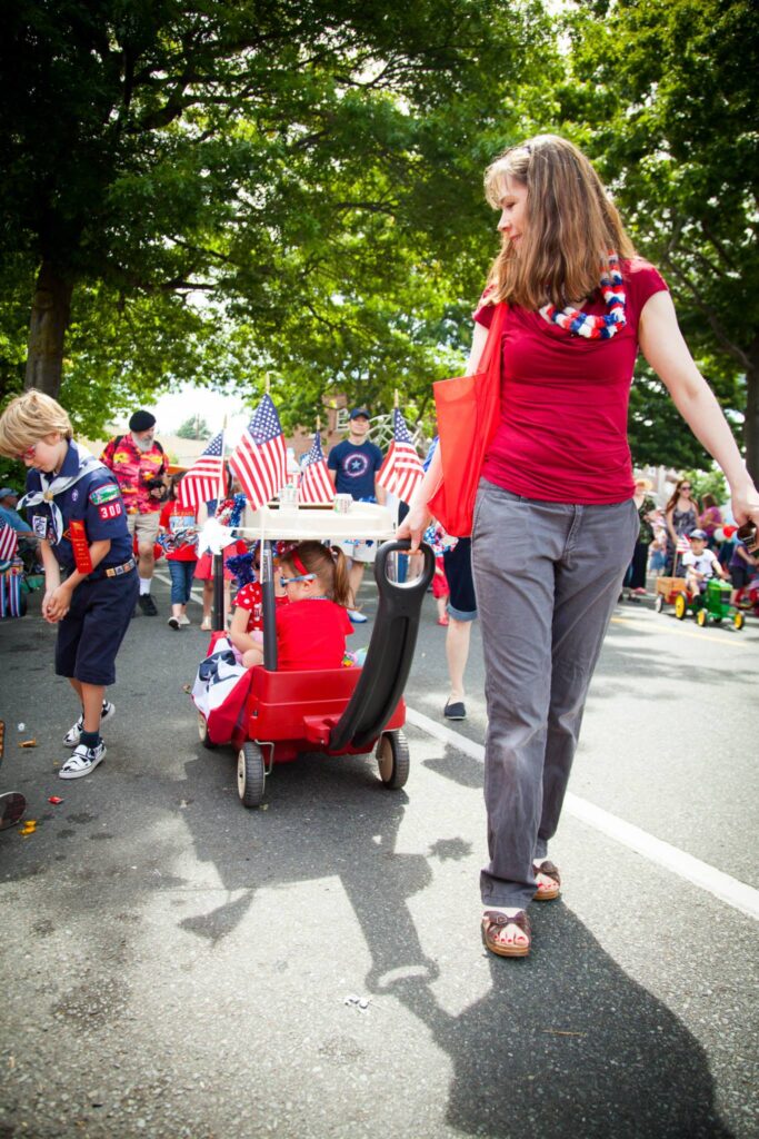 A person in a red shirt pushes a little red wagon full of cheers during a community parade with American flags.