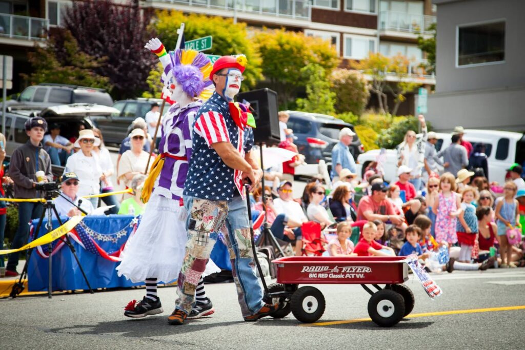Two performers in patriotic clown costumes push a red wagon through a sunny parade. Cheering spectators line the street in colorful summer attire.