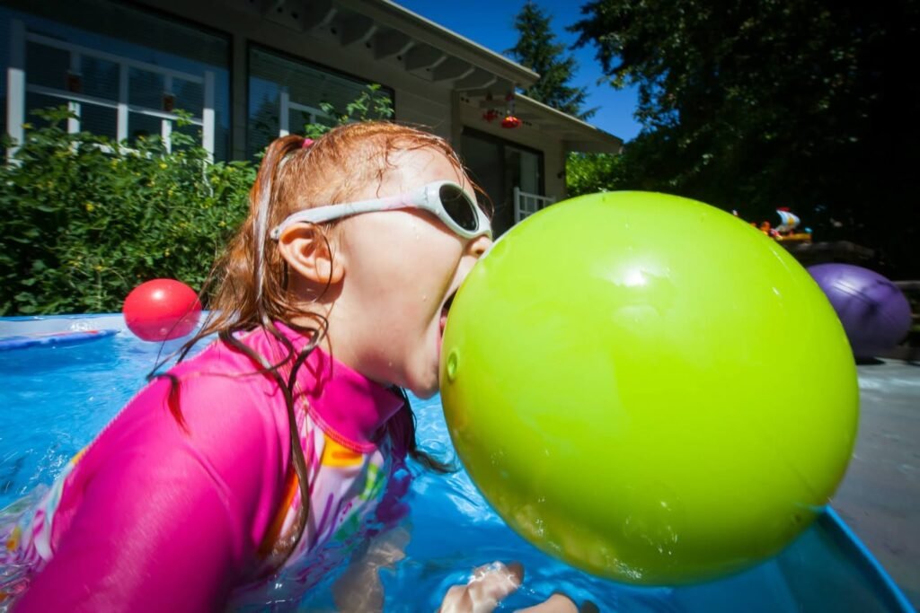 A child in a pink swimsuit blowing up a large green balloon in a swimming pool on a sunny day. The image captures a fun moment with vibrant colors and a playful atmosphere.