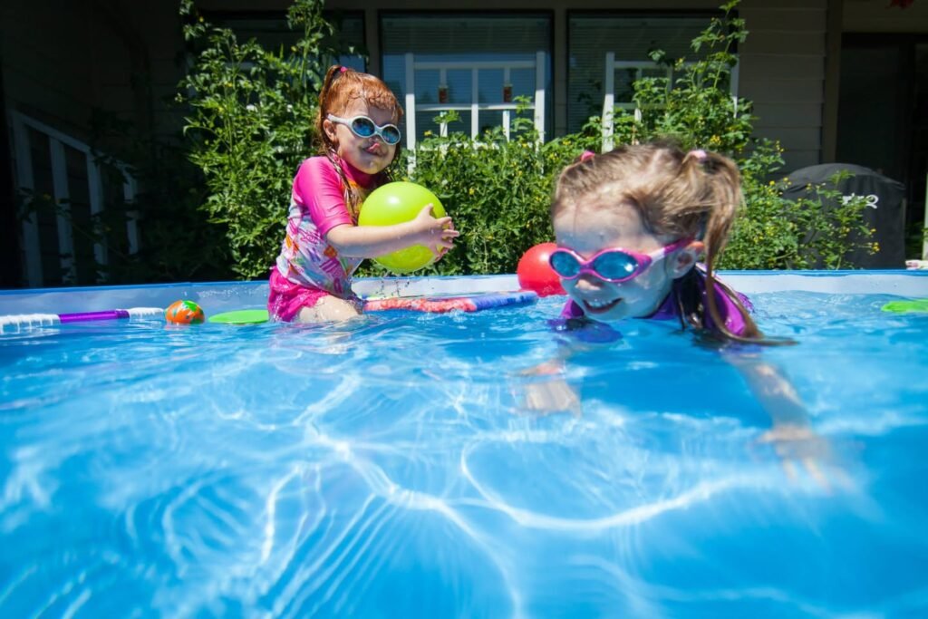 Two kids wearing swim goggles and pink rash guards play with colorful balls at the edge of a blue pool. Lush green bushes and a white railing are visible in the background.